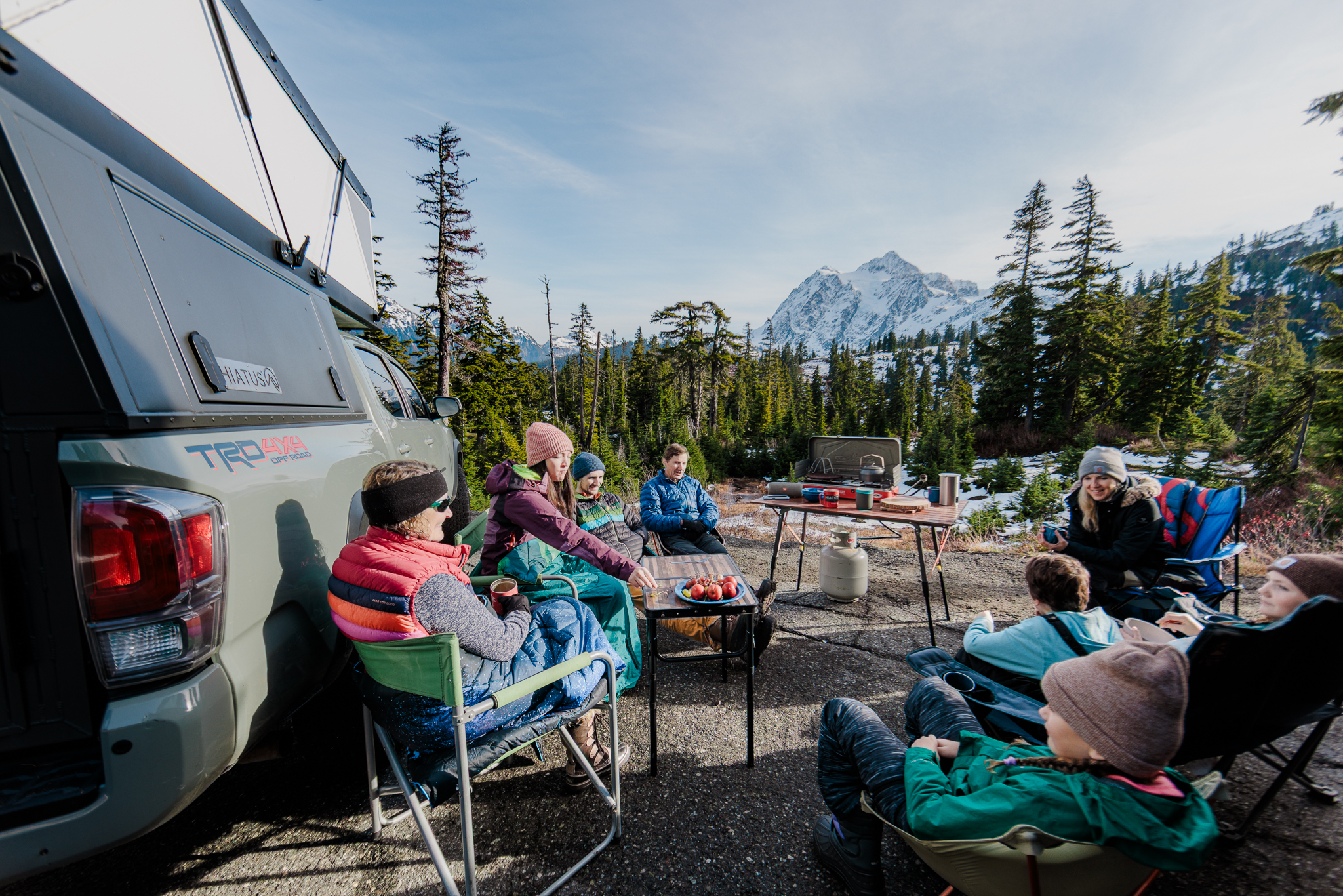 Family relaxing in camp chairs beside a Hiatus camper, enjoying hot cocoa during a winter truck camping trip.