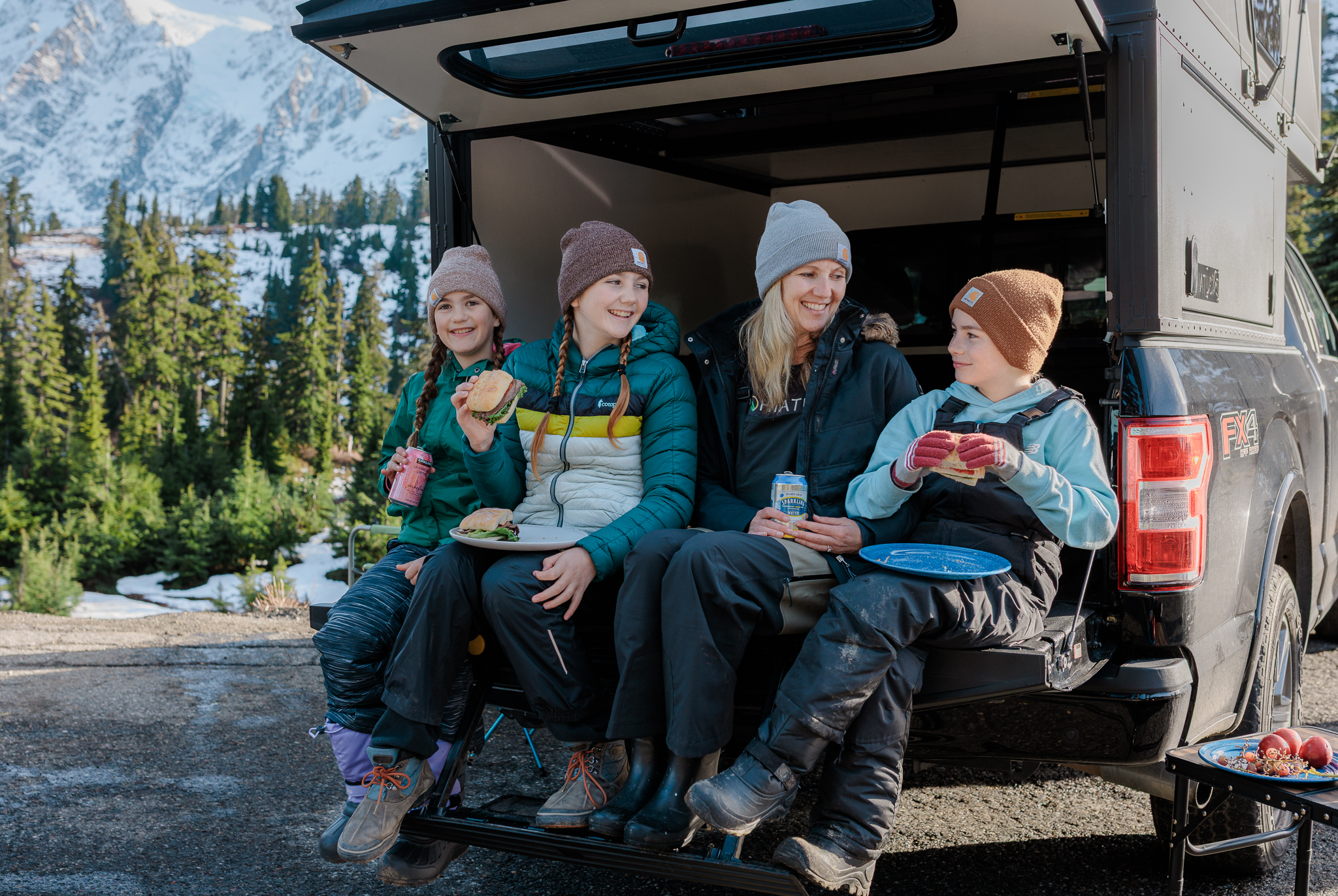 Family eating a tailgate meal at their Hiatus camper during a winter adventure with snowshoes and skis.